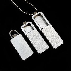 May include: Three rectangular metal containers with sliding lids, one featuring a wave pattern and another a honeycomb texture. The containers are silver-toned with a matte finish, and one is attached to a silver ball chain necklace.