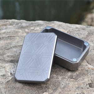 Könnte beinhalten: Rechteckige, silberfarbene Metallbox mit geometrischem Muster auf dem Deckel. Der Deckel ist leicht geöffnet und gibt den Blick ins Innere frei. Die Box steht auf einer Felsfläche. Minimalistisches Design.