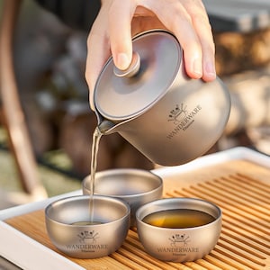 Könnte beinhalten: Eine matte graue Teekanne aus Titan mit Deckel und zwei passenden kleinen Tassen. Die Teekanne trägt den Schriftzug WANDERWARE Titanium und gießt Flüssigkeit in eine Tasse. Jede Tasse zeigt zudem das WANDERWARE Titanium Logo auf der Vorderseite.