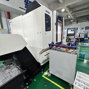 May include: A large, white and black CNC machine with the text "CMX 500 V" on the side. The machine is in a factory setting with a green floor and a metal shavings collection area.