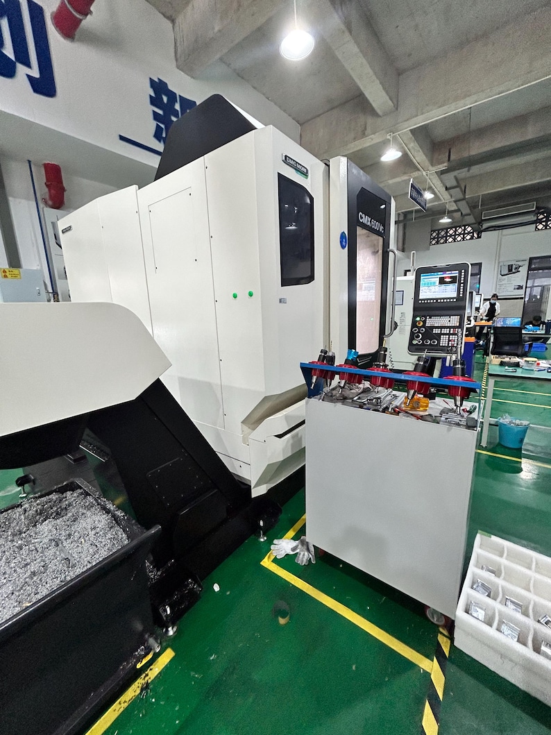 May include: A large, white and black CNC machine with the text "CMX 500 V" on the side. The machine is in a factory setting with a green floor and a metal shavings collection area.
