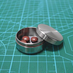 May include: A small, open stainless steel pill container with a matching lid. The container holds several brown pills. The lid is slightly ajar, revealing the contents. The container sits on a green surface.