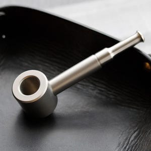 May include: A metallic smoking pipe with a cylindrical bowl and a long, straight stem. The pipe features a smooth, matte silver-toned finish throughout its entire structure, including the circular opening of the bowl and the flared mouthpiece.