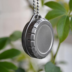 Puede incluir: Un colgante redondo plateado con un borde texturizado y un centro liso, suspendido de una cadena de bolas. El colgante es metálico y tiene un diseño utilitario. El fondo presenta hojas verdes y blancas borrosas.