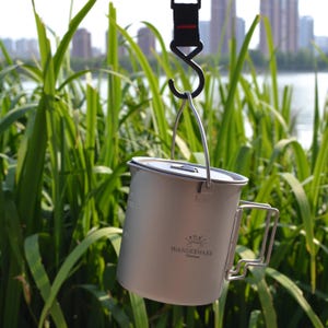 Puede incluir: Una taza de camping plateada con asa y tapa cuelga de un gancho negro. La taza tiene la inscripción "WANDERWARE Titanium". El fondo muestra hierba alta verde y un lago. Ideal para actividades al aire libre.