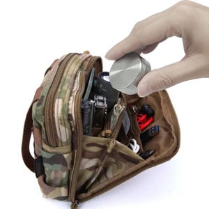 May include: A camouflage-patterned utility pouch with multiple compartments, filled with various items. A silver, circular object is held above the pouch. The pouch has a zipper closure and a brown interior.