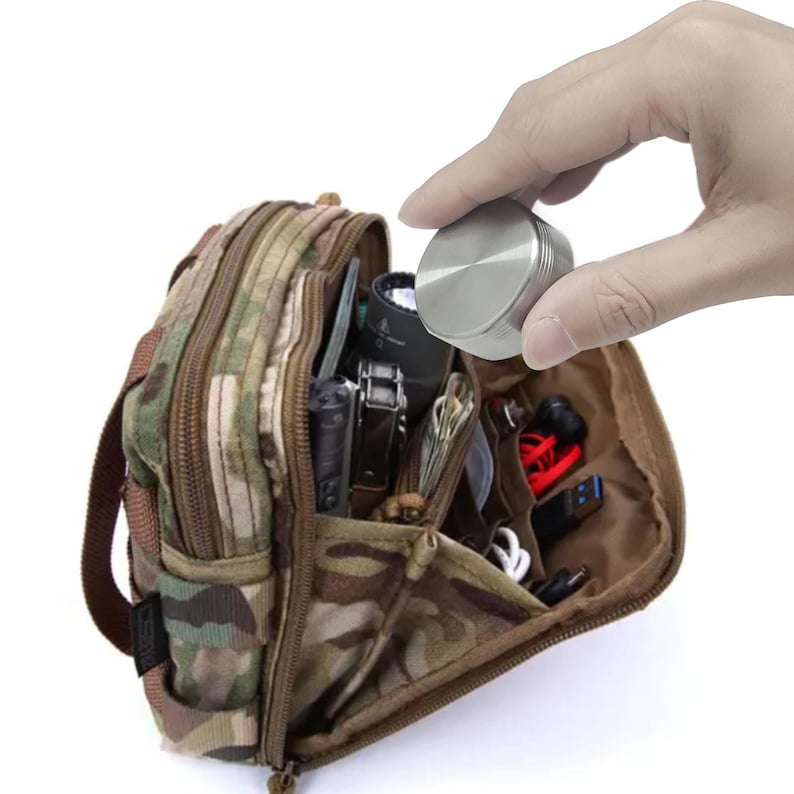 May include: A camouflage-patterned utility pouch with multiple compartments, filled with various items. A silver, circular object is held above the pouch. The pouch has a zipper closure and a brown interior.