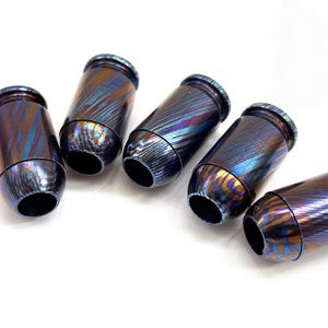 Puede incluir: Cinco objetos metálicos cilíndricos que se asemejan a casquillos de bala, cada uno con un acabado iridiscente con patrones de remolinos en tonos azul profundo, púrpura y cobre. Las superficies son pulidas y reflectantes con textura metálica.