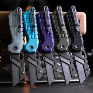 Könnte beinhalten: Fünf klappbare Gebrauchsmesser mit strukturierten Griffen in durchscheinendem Weiß, leuchtendem Blau, Violett, Olivgrün und Schwarz. Jedes Messer verfügt über eine dunkelgraue Metallklinge, einen Taschenclip und das North Mountain Blade Logo.