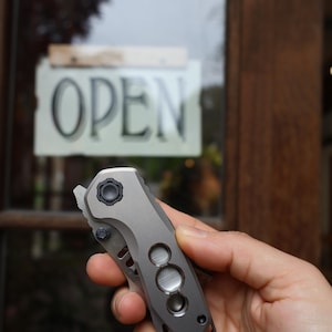 May include: A compact folding pocket knife with a matte grey titanium handle featuring a prominent circular pivot screw, a textured thumb stud, and three distinct circular cutouts along the side of the frame, held in a closed position.