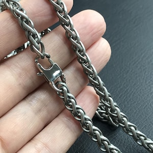以下が含まれることがあります： 磨き上げられた金属仕上げと安全なロブスタークラスプを備えた、シルバー調のウィートチェーンネックレス。チェーンは連動する楕円形のリンクで構成されており、ピースの全長にわたって編み込まれたような質感のある外観を作り出しています。