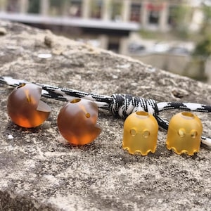 Op de afbeelding: Vier doorschijnende oortelefoontips in tinten amber en geel. Twee zijn gevormd als Pac-Man met een hap eruit, en twee zijn spookvormig. Een zwart-wit koord staat op de achtergrond.