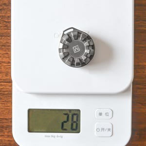 May include: A small, metallic, gear-shaped object with a 'W' logo is centered on a white digital scale. The scale displays the number '28'. The scale has a maximum capacity of 5 kg and a precision of 1g.