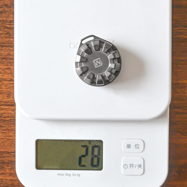 May include: A small, metallic, gear-shaped object with a 'W' logo is centered on a white digital scale. The scale displays the number '28'. The scale has a maximum capacity of 5 kg and a precision of 1g.