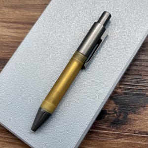 May include: A retractable ballpoint pen featuring a dark metallic grey cap and tip, paired with a translucent, frosted amber-coloured barrel. The pen includes a matching dark metallic grey pocket clip and a push-button mechanism at the top end.