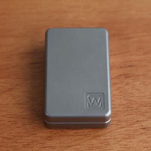 Puede incluir: Una caja rectangular de metal gris con esquinas redondeadas. La caja tiene un pequeño diseño en relieve en la esquina inferior derecha. Está sobre una superficie de madera.