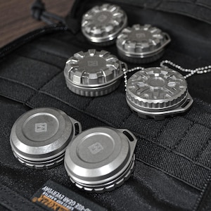 May include: A collection of small, metallic, round containers with intricate designs. Some have a loop for attachment, and one is attached to a chain. The containers are arranged on a black, textured surface.