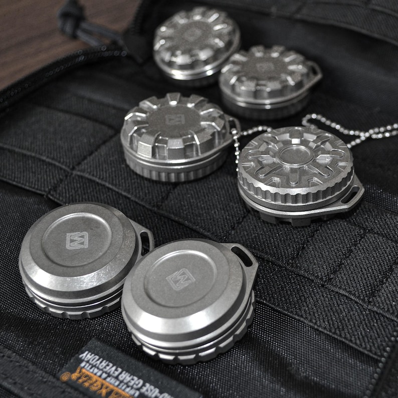 May include: A collection of small, metallic, round containers with intricate designs. Some have a loop for attachment, and one is attached to a chain. The containers are arranged on a black, textured surface.