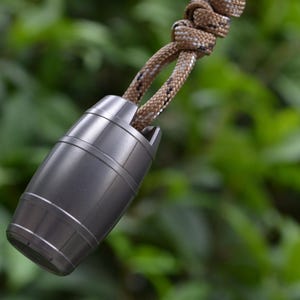 May include: A metallic gray barrel-shaped bead with a smooth finish and horizontal grooves, attached to a thick, braided tan and brown cord that features a prominent knot at the top, creating a functional and durable accessory component.