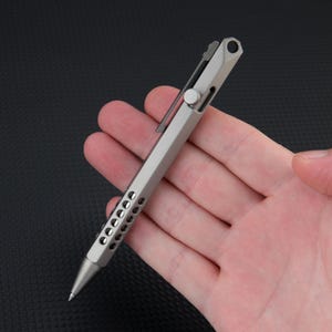 May include: A metallic gray bolt-action pen with a hexagonal barrel, featuring a series of circular cutouts along the lower grip section, a side-mounted sliding mechanism, a pocket clip, and a small circular hole at the top end of the pen body.