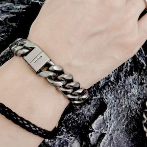 Peut inclure: Un bracelet chaîne argenté avec un design à maillons larges. Le bracelet est doté d'un fermoir rectangulaire avec un détail gravé subtil. Un bracelet en cuir tressé noir est également visible.