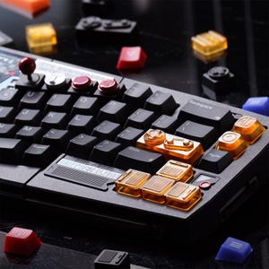 Könnte beinhalten: Eine schwarze mechanische Tastatur mit individuellen Tastenkappen in verschiedenen Farben, darunter Orange, Rot und Blau. Die Tastatur hat einen Joystick und mehrere benutzerdefinierte Tasten. Der Text "2015-2025" ist sichtbar.