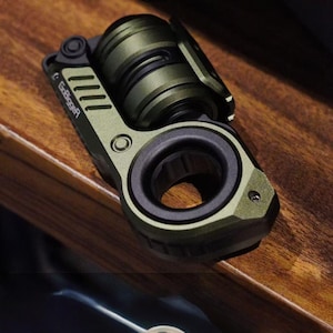 May include: An olive green multi-tool with a circular opening and a cylindrical component. The tool has a textured surface and the text "GoBigger" is visible. The tool is set against a dark wood grain background.