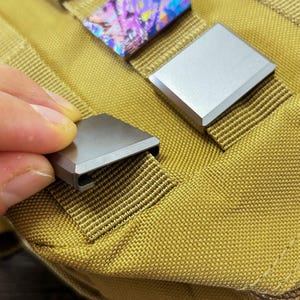 Può includere: Primo piano di tre fibbie quadrate in metallo argentato e iridescente. Una fibbia viene inserita in una cinghia di tessuto color cachi. Le fibbie sono progettate per fissare cinghie o fettucce.