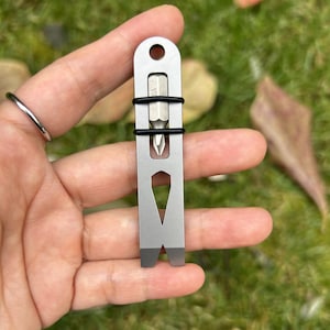 May include: A small, silver multi-tool with a screwdriver bit and pry bar end. The tool has a hole at the top for attaching to a keyring. Two black rubber bands secure the screwdriver bit. The tool is held in a hand against a blurred green background.