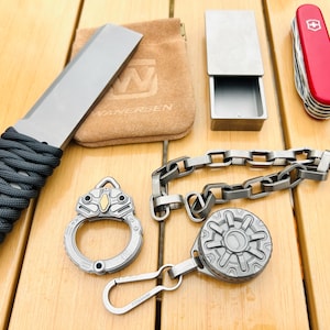 May include: An assortment of metallic gray EDC (Everyday Carry) items, including a knife with a wrapped handle, a small box, a chain, a keychain with a carabiner, and a multi-tool. A small brown pouch is also present.