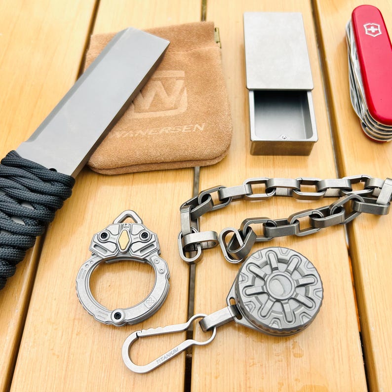 May include: An assortment of metallic gray EDC (Everyday Carry) items, including a knife with a wrapped handle, a small box, a chain, a keychain with a carabiner, and a multi-tool. A small brown pouch is also present.
