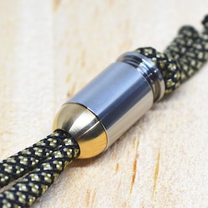 May include: Close-up of a metal clasp with a cylindrical silver body and gold-colored ends. The clasp is attached to a black and gold braided cord, likely for a necklace or bracelet. The background is a light-colored wooden surface.