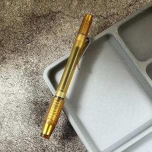 May include: A gold-coloured pen with a textured grip and a silver clip rests on a grey tray. The pen's body has a translucent appearance, and the tip and end are solid gold. The tray has multiple compartments, suggesting organisation.