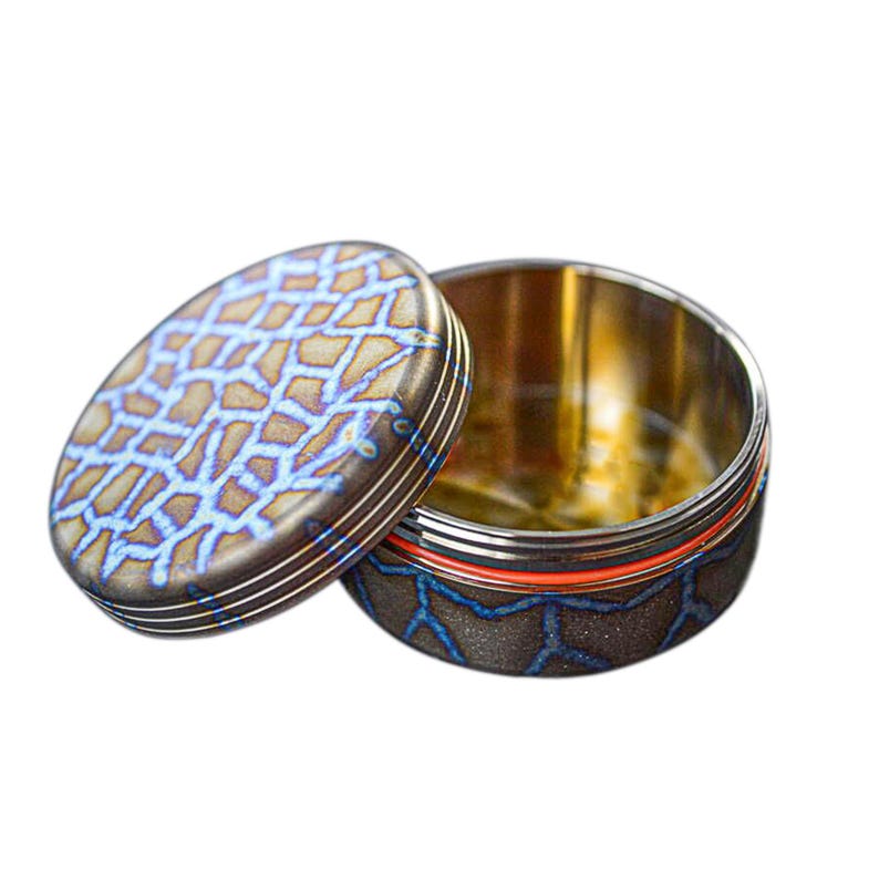 May include: A small, round container with a lid. The lid and base have a dark, textured pattern with blue accents. The interior is gold-colored, and a red band encircles the base. The lid is slightly ajar, revealing the inside.