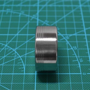May include: A cylindrical, stainless steel ring with etched horizontal lines. The ring is highly polished, reflecting the surrounding environment. The design features a series of parallel grooves near the top and bottom edges.