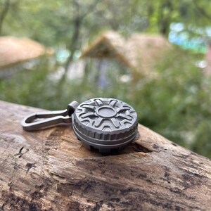 May include: A small, gray, circular keychain accessory with a detailed geometric design on top. It has a matching gray clip attached. The item is resting on a piece of wood, with a blurred green background.