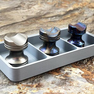 May include: Three metal stands with concave tops arranged in a light gray rectangular tray. The first stand is matte silver, while the second and third stands feature iridescent, heat-treated finishes with swirling blue, purple, and bronze tones.