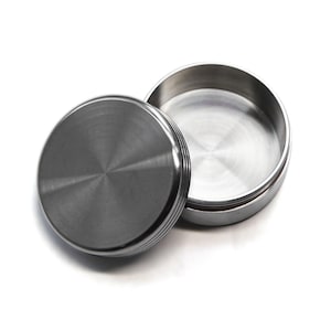 May include: A stainless steel container with a lid. The lid is off to the side, revealing the inside of the container. The container and lid have a brushed metal finish. The lid has a series of horizontal ridges around the edge.