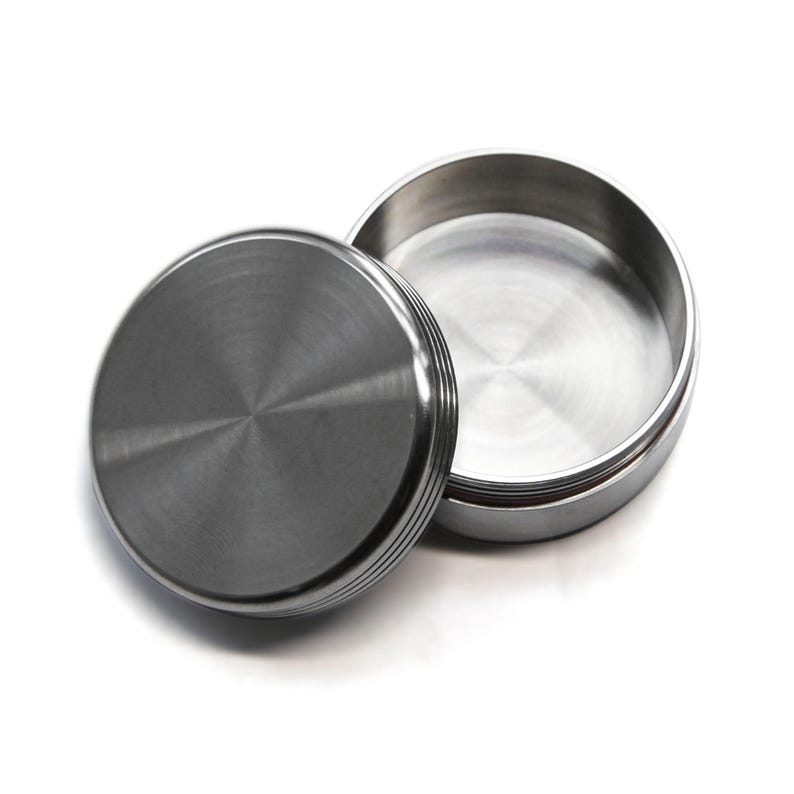 May include: A stainless steel container with a lid. The lid is off to the side, revealing the inside of the container. The container and lid have a brushed metal finish. The lid has a series of horizontal ridges around the edge.