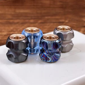 Op de afbeelding: Vier cilindervormige kralen met een messingkleurige kern en een gefacetteerde buitenkant. De kralen hebben verschillende afwerkingen: één matzwart, één doorschijnend blauw, één met een veelkleurig iriserend patroon en één matgrijze metallic afwerking.