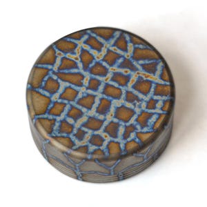 May include: A round, decorative object with a textured surface. The top features a brown and blue geometric pattern. The sides have a similar design. The object is set against a white background.