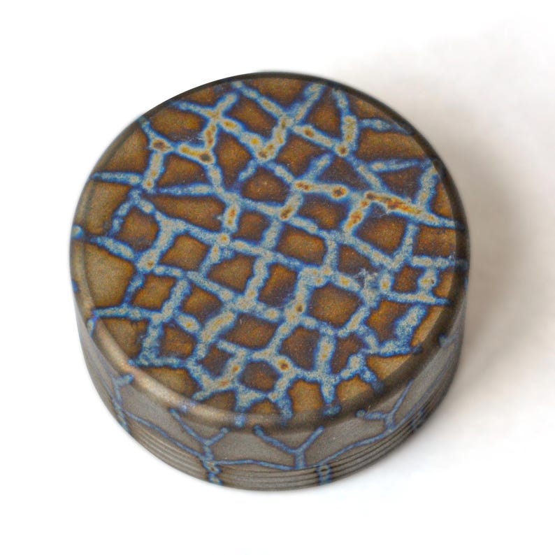 May include: A round, decorative object with a textured surface. The top features a brown and blue geometric pattern. The sides have a similar design. The object is set against a white background.
