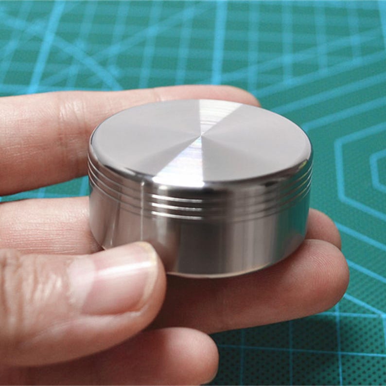 May include: A small, cylindrical, brushed metal object with a ridged side and a flat, circular top. The top has a radial pattern emanating from the center. The object is held in a person's hand against a green background.