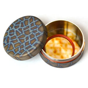 May include: An open, oval-shaped container with a lid. The lid and base have a bronze finish with a blue, mosaic-like pattern. Inside, there are two circular, light-colored objects and a red ring.
