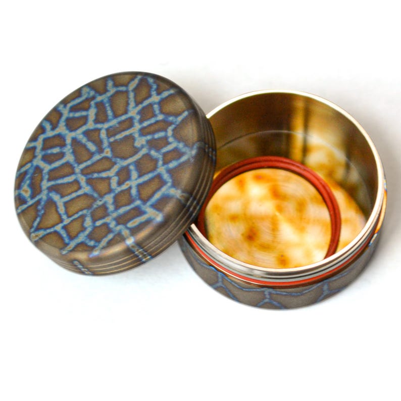 May include: An open, oval-shaped container with a lid. The lid and base have a bronze finish with a blue, mosaic-like pattern. Inside, there are two circular, light-colored objects and a red ring.