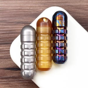 May include: Three small, cylindrical containers with textured surfaces. One is silver, one is amber, and the third is blue and purple. The containers are arranged on a white surface against a wooden background. The containers appear to be made of metal.