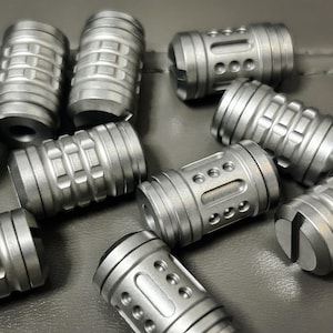 May include: A collection of small, cylindrical metal objects with various surface designs. The objects are silver in color and appear to be made of a durable metal. Some have grooves, while others have holes or cut-out patterns. The objects are scattered.