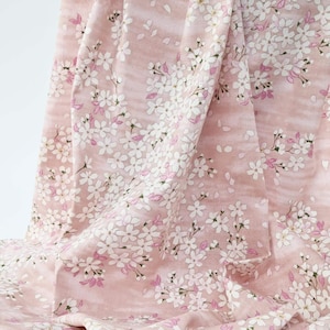 May include: A draped fabric featuring a delicate floral pattern. The design showcases white cherry blossoms with pink accents against a soft pink background. The fabric's folds create a sense of depth and texture, ideal for textiles.
