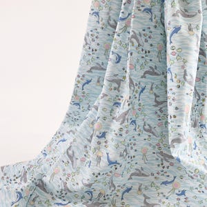 May include: A light blue fabric featuring a repeating pattern of grey whales, blue dolphins, pink jellyfish, and small colorful fish. The design is set against a background of wavy white lines, creating an underwater scene across the draped textile.