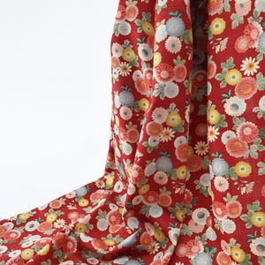 May include: Red fabric with a repeating floral pattern. The flowers are in shades of pink, yellow, blue, and white. The fabric is draped, showing the texture and flow of the material. The background is white.